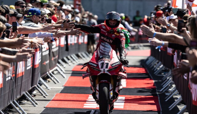 Motorcycle racer in red gear high-fives fans along a red-carpet finish line, crowds on both sides reaching out to celebrate a victory.