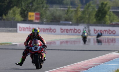 MotoGP rider in red leathers leaning into a corner, leading on a red-and-white bike with the number 5; Brembo signs in the background.