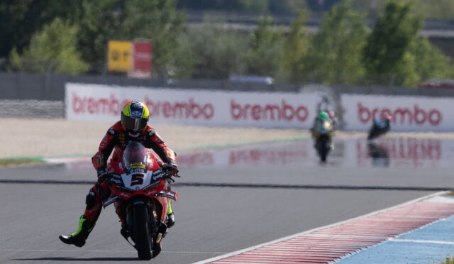 MotoGP rider in red leathers leaning into a corner, leading on a red-and-white bike with the number 5; Brembo signs in the background.