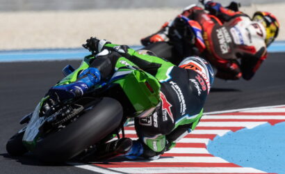 Motorcycle racers lean into a sharp turn on a race track, green Kawasaki bike in front with blue accents and other riders trailing behind on red-and-white striped curbs.