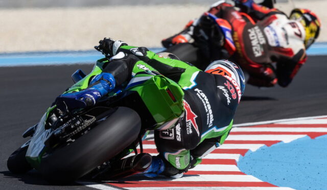 Motorcycle racers lean into a sharp turn on a race track, green Kawasaki bike in front with blue accents and other riders trailing behind on red-and-white striped curbs.