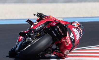 Motorcycle racer in red leathers leaning into a tight turn on a race track next to red and white curb marks.