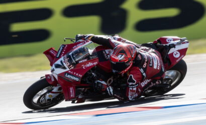 Professional motorcycle racer in a red suit leaning into a turn on a red race bike with sponsor logos visible on the bodywork and helmet.