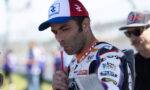 Racer in a white-red-blue cap and racing suit drinks from a red straw before a race.