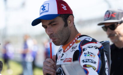 Racer in a white-red-blue cap and racing suit drinks from a red straw before a race.