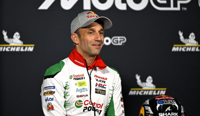 MotoGP rider in a white-green-red racing suit with a gray Red Bull cap, standing on a stage with Michelin logos in the background, helmet nearby.