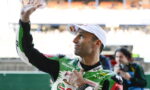 Racing rider in a green and white suit waves to the crowd, wearing a white cap at the track.