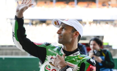 Racing rider in a green and white suit waves to the crowd, wearing a white cap at the track.