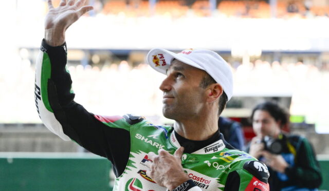 Racing rider in a green and white suit waves to the crowd, wearing a white cap at the track.