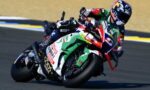 Motorcycle racer leaning into a turn on a racetrack, with a colorful sponsor-labeled bike visible in the foreground.