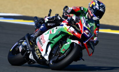 Motorcycle racer leaning into a turn on a racetrack, with a colorful sponsor-labeled bike visible in the foreground.