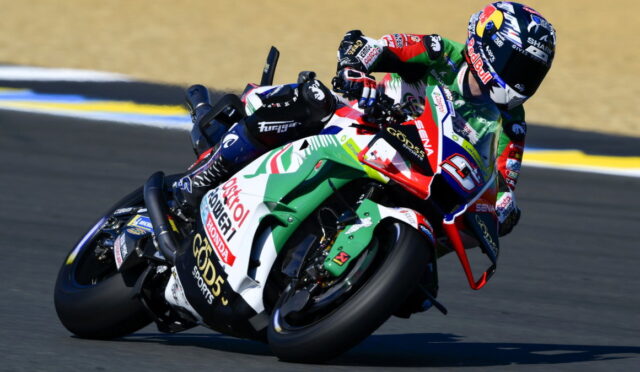 Motorcycle racer leaning into a turn on a racetrack, with a colorful sponsor-labeled bike visible in the foreground.