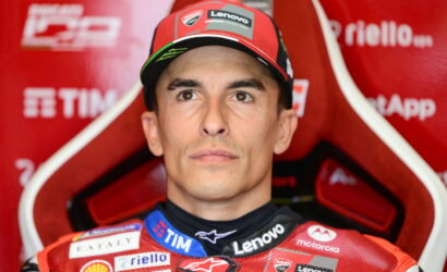 Portrait of a motorcycle racer in a red racing suit and cap, sitting in a red team chair with sponsor logos visible (Lenovo, TIM).