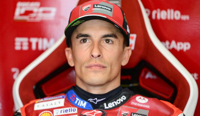 Portrait of a motorcycle racer in a red racing suit and cap, sitting in a red team chair with sponsor logos visible (Lenovo, TIM).
