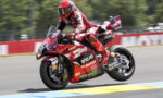 Rider in red racing suit on a red Ducati sport bike leans into a corner on a race track, speed lines blurred in the background.