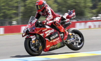 Rider in red racing suit on a red Ducati sport bike leans into a corner on a race track, speed lines blurred in the background.