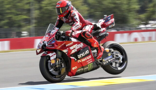 Rider in red racing suit on a red Ducati sport bike leans into a corner on a race track, speed lines blurred in the background.