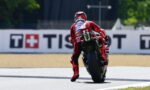 MotoGP rider in red racing suit leans into a turn on a race track, rear wheel skidding slightly, sponsor logos visible on suit and bike.