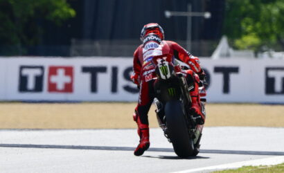 MotoGP rider in red racing suit leans into a turn on a race track, rear wheel skidding slightly, sponsor logos visible on suit and bike.