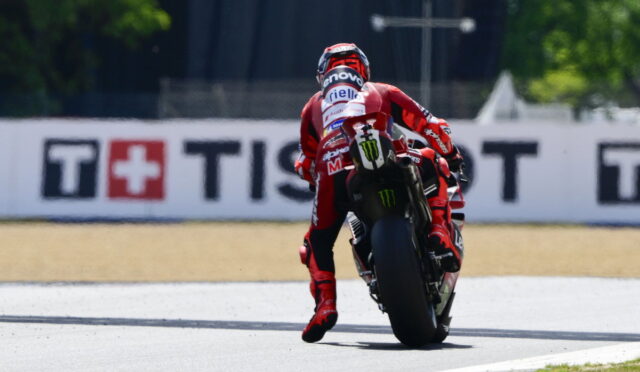 MotoGP rider in red racing suit leans into a turn on a race track, rear wheel skidding slightly, sponsor logos visible on suit and bike.