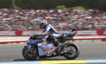 Blue racing motorcycle with sponsor stickers speeds on track as a blurred stadium crowd watches in the background.