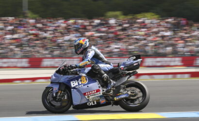 Blue racing motorcycle with sponsor stickers speeds on track as a blurred stadium crowd watches in the background.