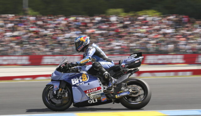 Blue racing motorcycle with sponsor stickers speeds on track as a blurred stadium crowd watches in the background.