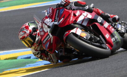 Motorcycle racer in red leans into a sharp turn on a race track, sponsor logos visible on suit and bike, blue-yellow-green curbs in the background.