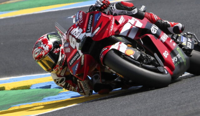 Motorcycle racer in red leans into a sharp turn on a race track, sponsor logos visible on suit and bike, blue-yellow-green curbs in the background.