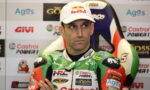 Racing rider wearing a Red Bull cap sits in a press area with sponsor logos in the background, green Castrol racing suit visible.