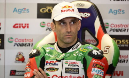 Racing rider wearing a Red Bull cap sits in a press area with sponsor logos in the background, green Castrol racing suit visible.