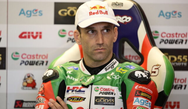 Racing rider wearing a Red Bull cap sits in a press area with sponsor logos in the background, green Castrol racing suit visible.