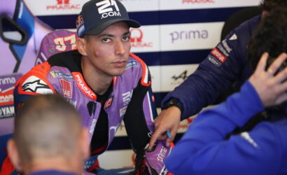 MotoGP rider in a purple racing suit sits in the pit area as a team member in blue writes on his forearm, with sponsor banners in the background.