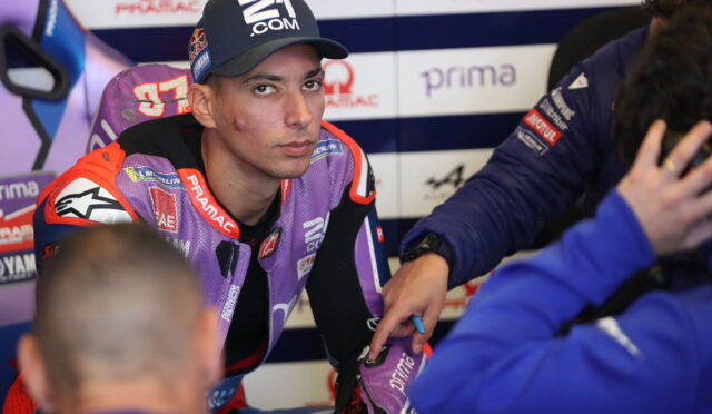 MotoGP rider in a purple racing suit sits in the pit area as a team member in blue writes on his forearm, with sponsor banners in the background.