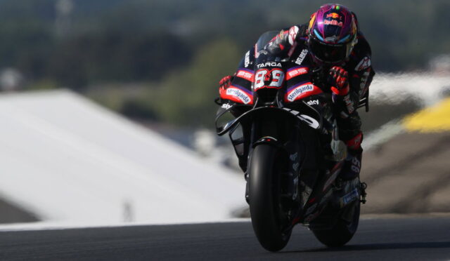 Racing motorcyclist in full gear leans into a turn on a track, colorful helmet and sponsor livery visible.