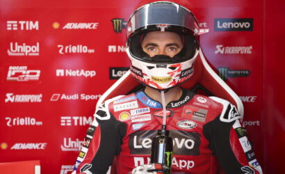 MotoGP racer in a red Lenovo racing suit sits in a red sponsor-filled backdrop, wearing a helmet and holding a water bottle.