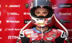 Motorcycle racer in red racing suit and helmet, facing the camera against a red sponsor backdrop