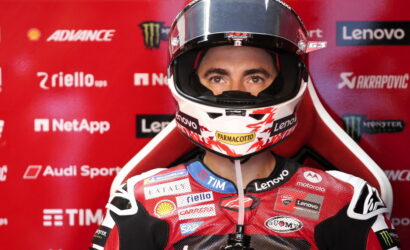 Motorcycle racer in red racing suit and helmet, facing the camera against a red sponsor backdrop
