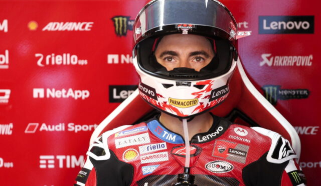 Motorcycle racer in red racing suit and helmet, facing the camera against a red sponsor backdrop
