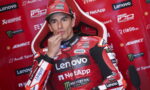 MotoGP rider in a red Lenovo NetApp racing suit sits in a sponsor backdrop, hand at his chin.