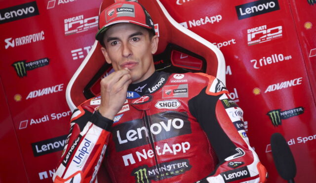 MotoGP rider in a red Lenovo NetApp racing suit sits in a sponsor backdrop, hand at his chin.