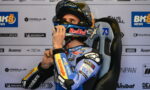 Moto racer in blue suit sits in a racing chair, adjusts helmet before race with sponsor logos in the background.