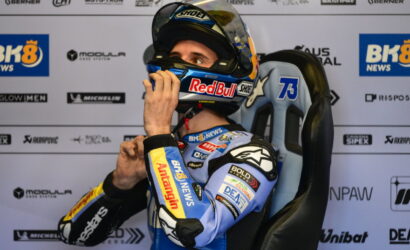 Moto racer in blue suit sits in a racing chair, adjusts helmet before race with sponsor logos in the background.