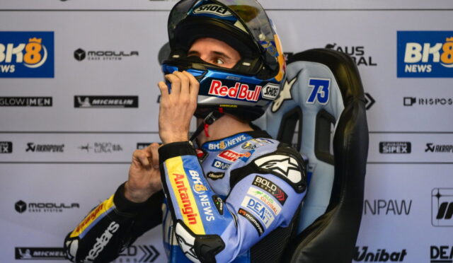 Moto racer in blue suit sits in a racing chair, adjusts helmet before race with sponsor logos in the background.
