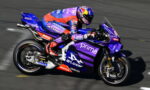 Rider in red and blue suit leans into a turn on a purple Yamaha race bike with Prima branding on a race track.