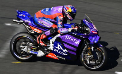 Rider in red and blue suit leans into a turn on a purple Yamaha race bike with Prima branding on a race track.
