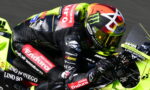 MotoGP rider in black and red leathers on a neon-yellow racing bike, leaning into a turn with a colorful helmet visible.