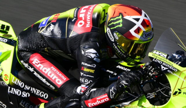 MotoGP rider in black and red leathers on a neon-yellow racing bike, leaning into a turn with a colorful helmet visible.