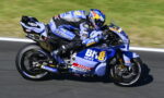 Blue and white racing motorcycle with rider in full gear leaning into a turn on a race track, sponsor logos visible.
