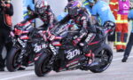 Two professional motorcycle racers lean into a turn on a race track, wearing full leathers and helmets; bikes are black with red/white decals and sponsor logos, with pit crew in the background.